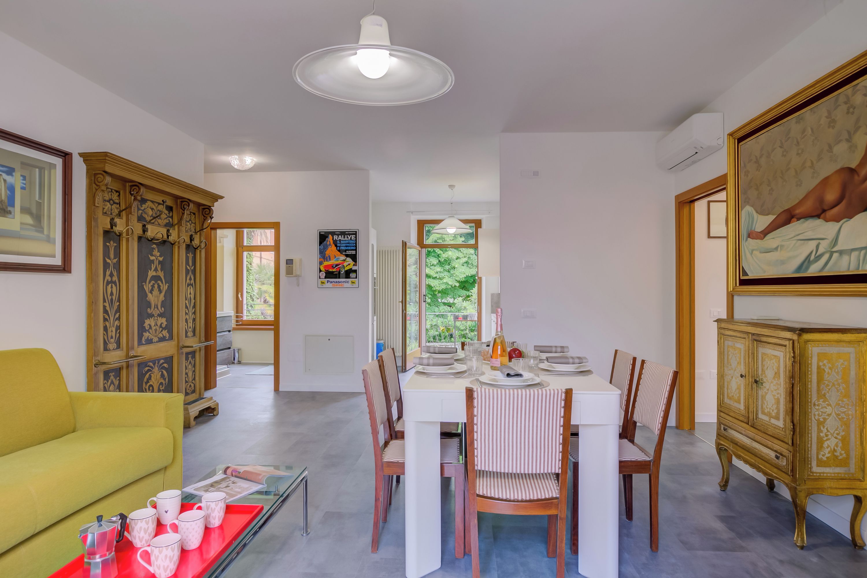 Ein Essbereich mit einem weißen Tisch, gedeckt für vier Personen, umgeben von Holzstühlen. Im Vordergrund ist eine gelbe Couch zu sehen, und auf dem Tisch steht ein roter Tablett mit Tassen und einer Karaffe.