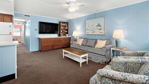 A living room with a sofa, a coffee table, a television, a bookshelf, and a painting on the wall. - Three Bedroom Oceanside Condo Steps from the Beach (Ocean City)