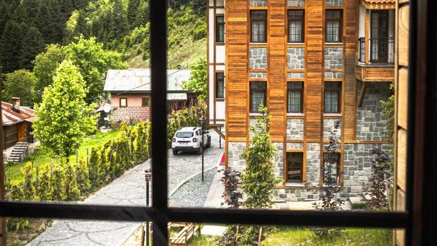 A view through a window of a building with a stone facade and wooden accents, surrounded by greenery and a hilly landscape.
