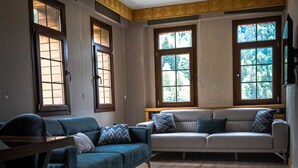 A living room with a blue sofa, a grey sofa, and a wooden coffee table. There are three windows with wooden frames, and a large rug on the floor.