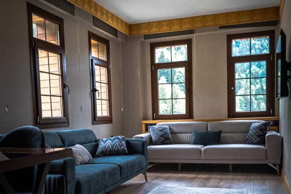 Sala de estar con un sofá azul, un sofá gris y una mesa de centro de madera. Hay tres ventanas con marcos de madera, y una alfombra grande en el suelo.