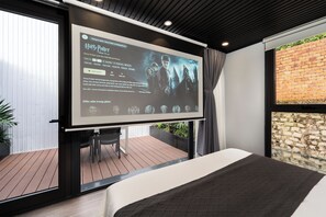 Una camera da letto con una grande TV a schermo piatto fissata al muro, un letto e vista su un pontile in legno e un muro di mattoni.