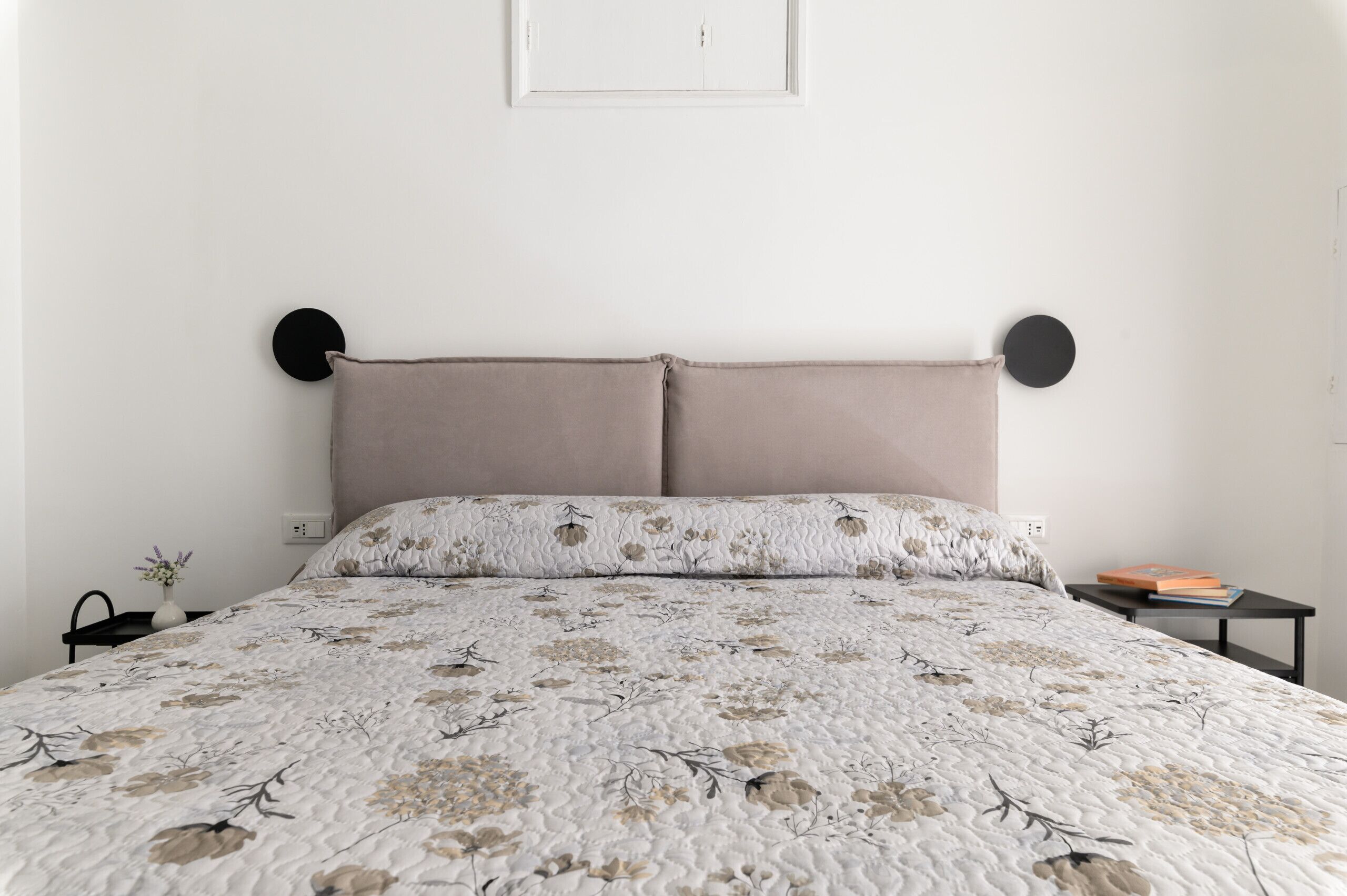 Una cama con colcha floral, una mesita de noche con un libro y un reloj en la pared.