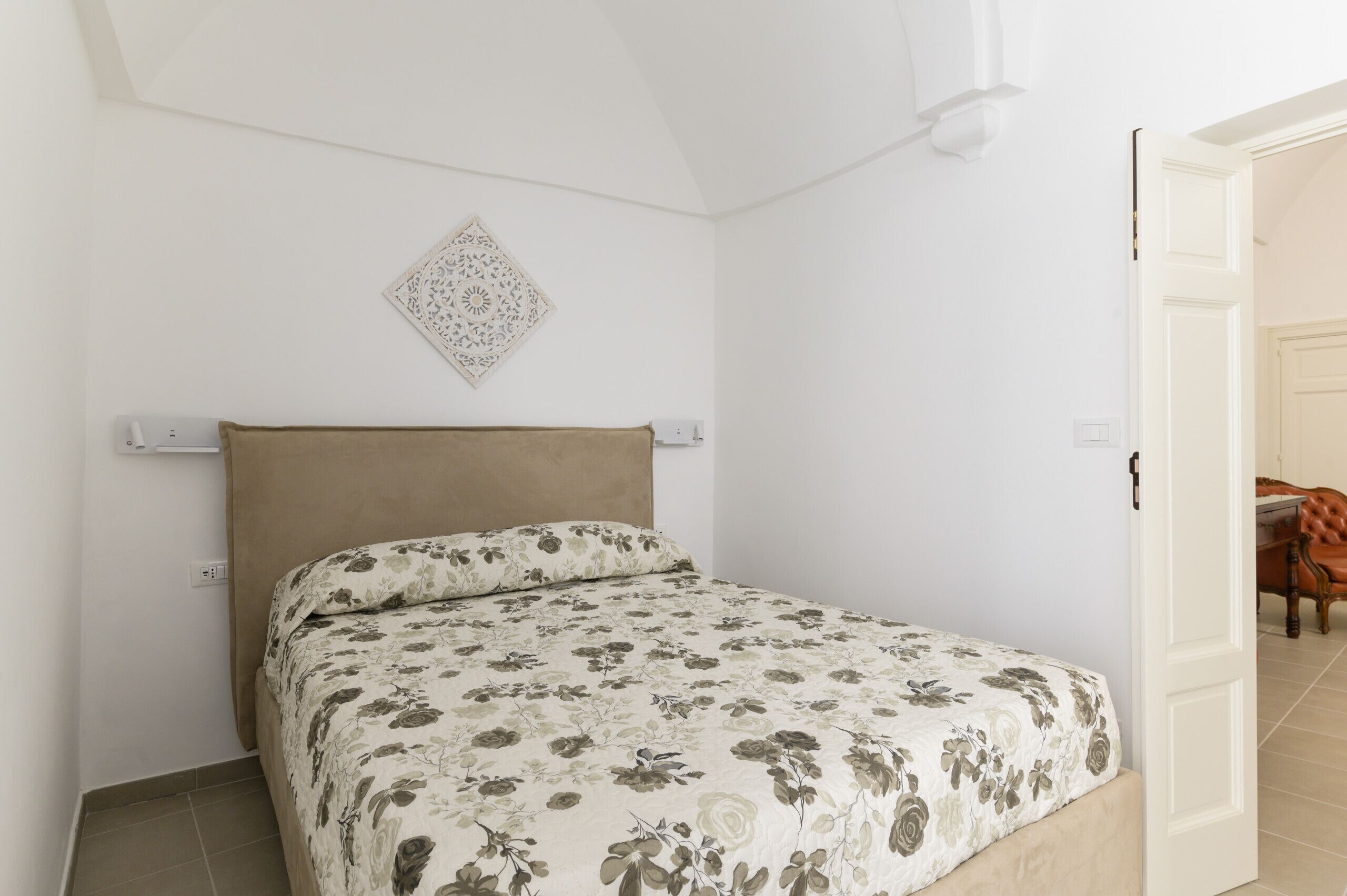 A bedroom with a bed, a headboard, a wall-mounted light fixture, and a door leading to another room.