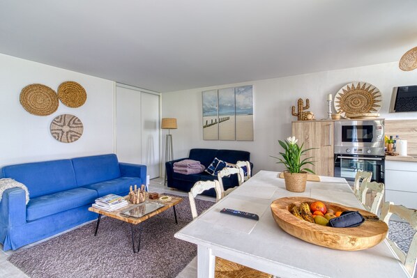 Un salón con un sofá azul, una mesa de centro de madera y una mesa de comedor con un frutero. Hay una zona de cocina con microondas y un televisor.