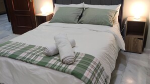 A neatly made bed with a plaid blanket and rolled towels, two bedside tables with lamps, and a wooden door.