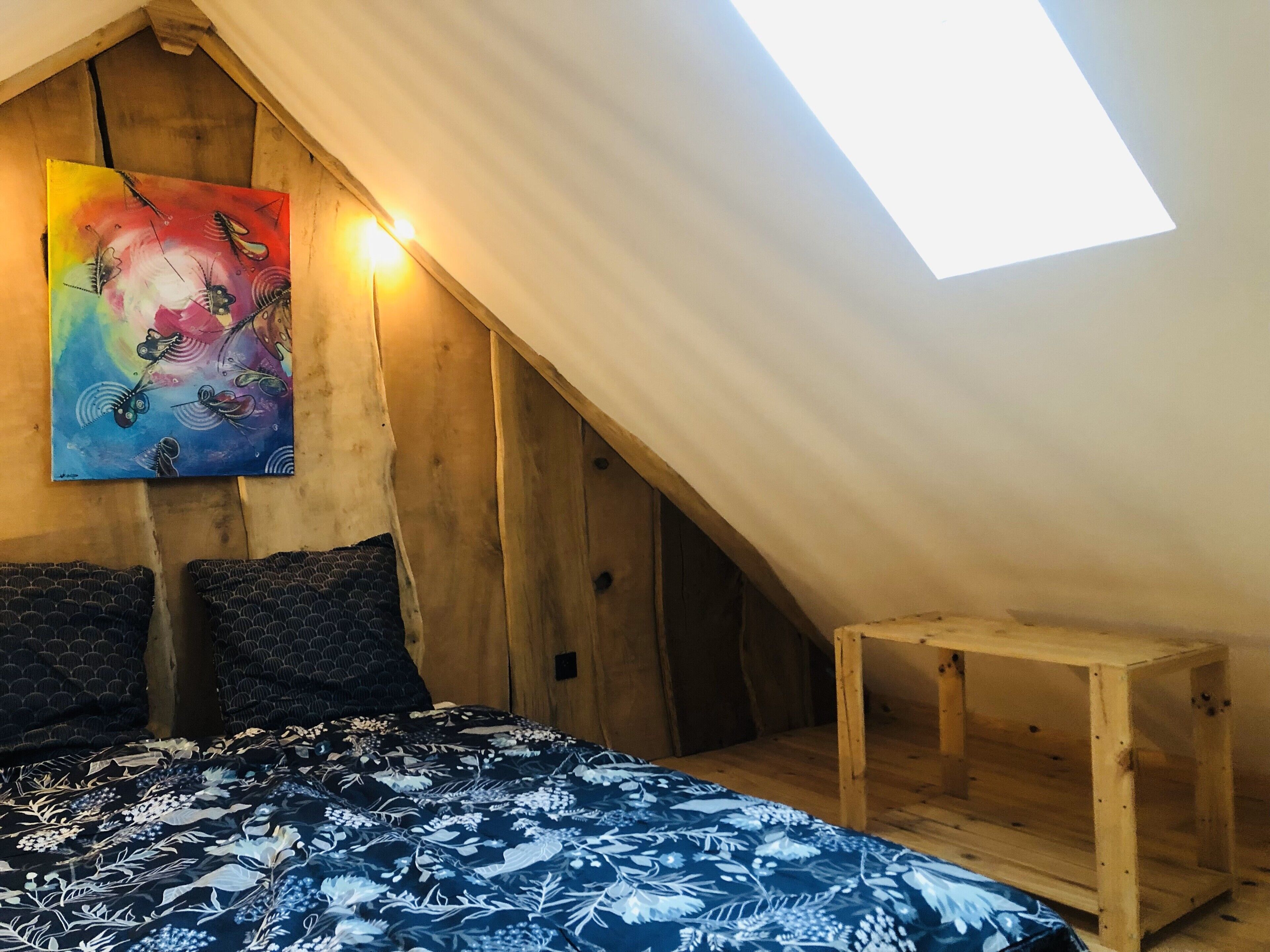 Une chambre avec un plafond en pente, un tableau, un lit avec une literie à motifs et une petite table en bois.