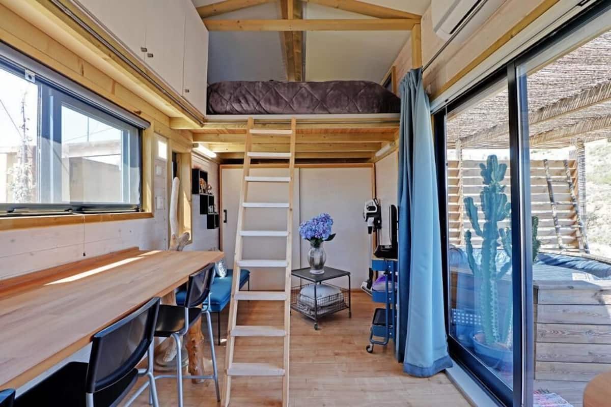 A compact interior with a bunk bed, a dining area with a wooden table and chairs, and a large sliding glass door leading to an outdoor area with a cactus.