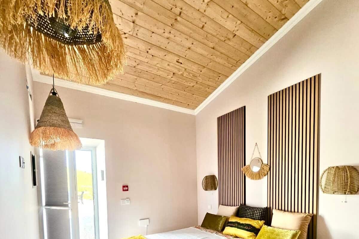 A bedroom with a wooden ceiling, a bed with a white and yellow bedspread, and a hanging light fixture.
