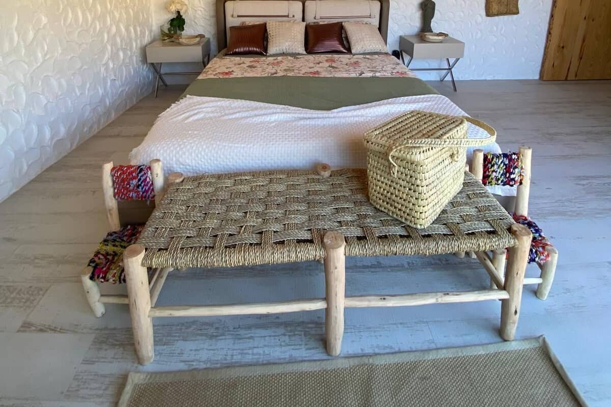 A bedroom with a bed, two nightstands, a woven bench, and a hanging basket.