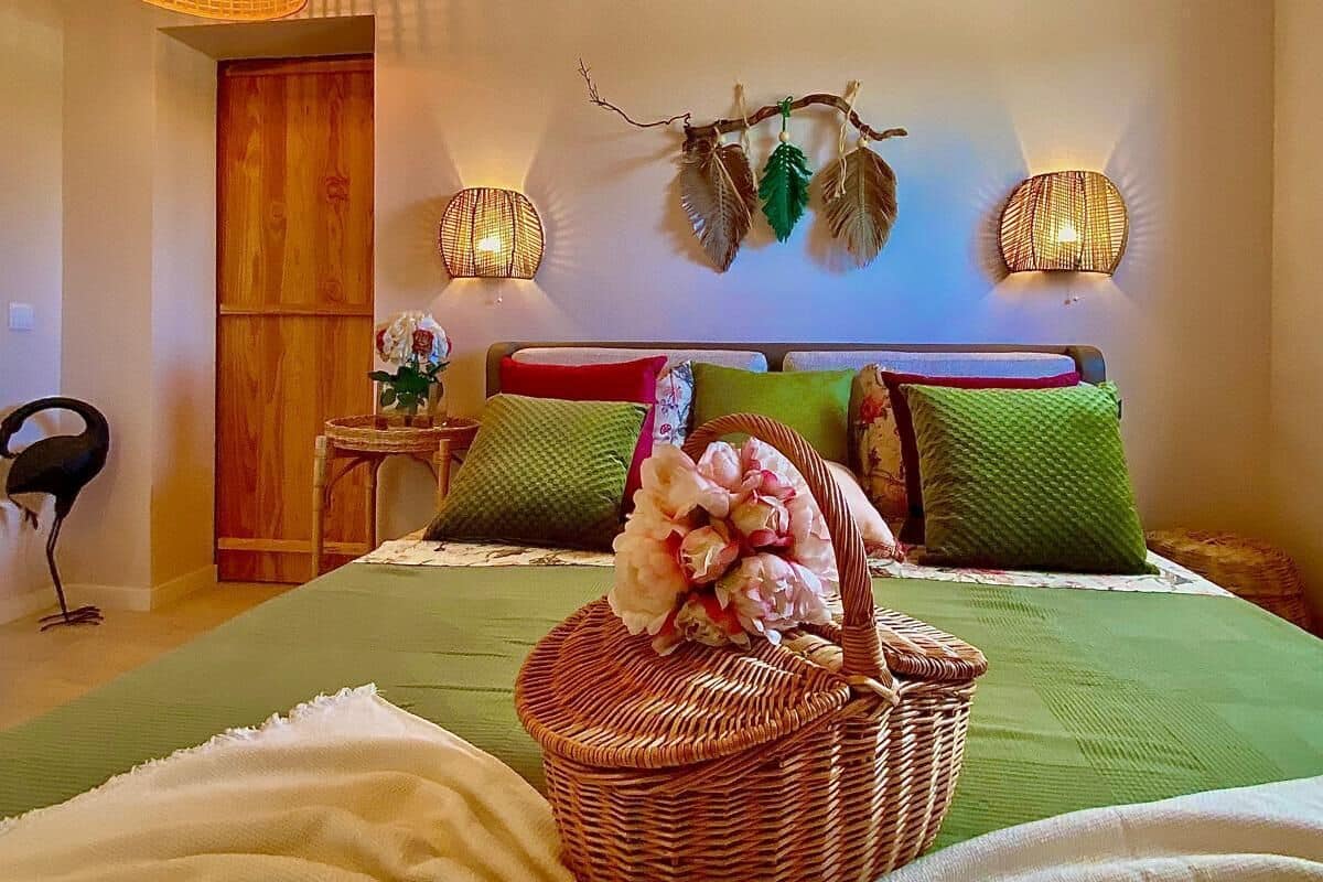 A bedroom with a bed, green bedding, a wicker basket with flowers, a wooden wardrobe, and wall-mounted lights.