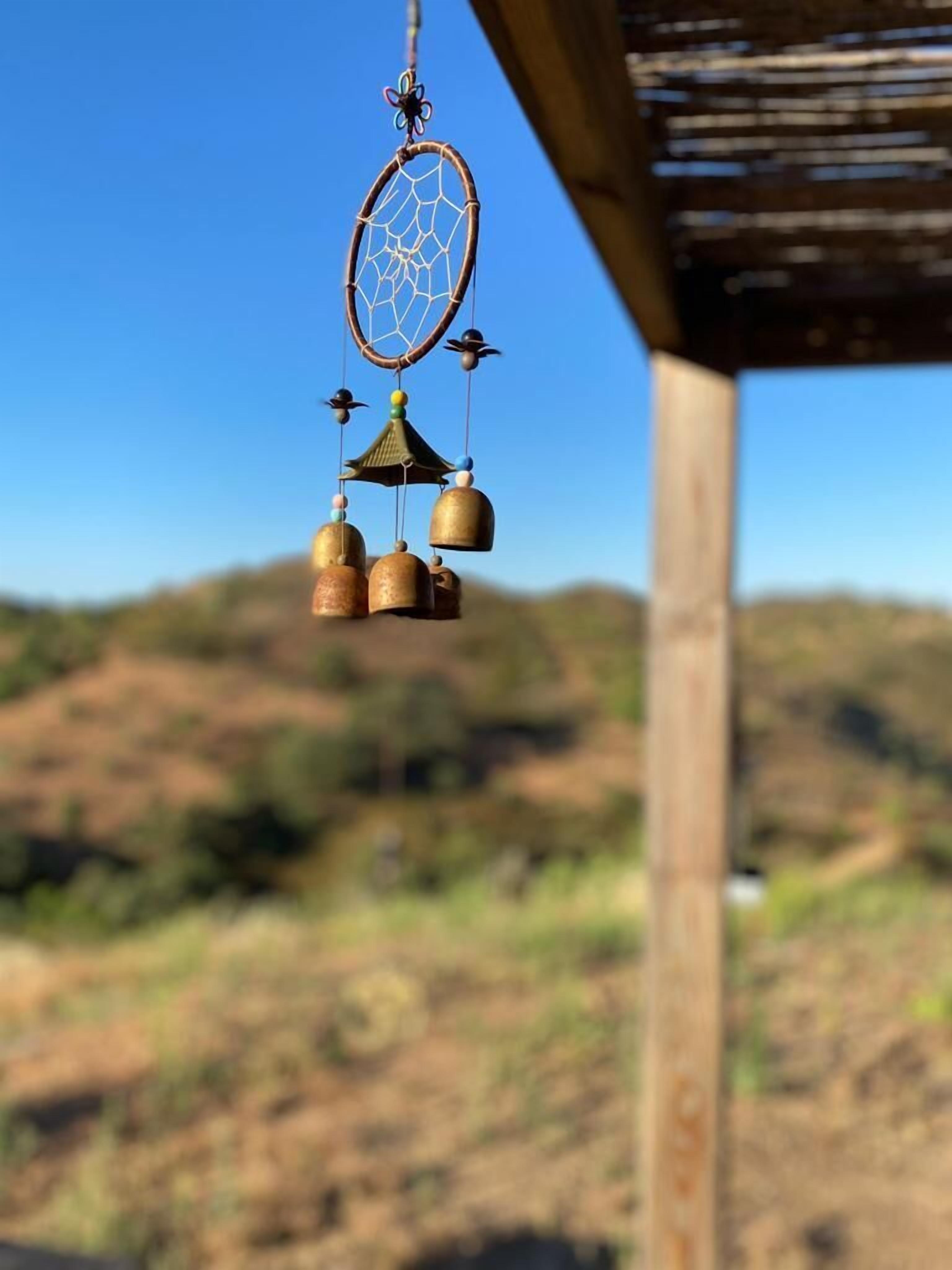 Un atrapasueños con cascabeles y un adorno colgante, frente a un cielo despejado, con un paisaje borroso al fondo.