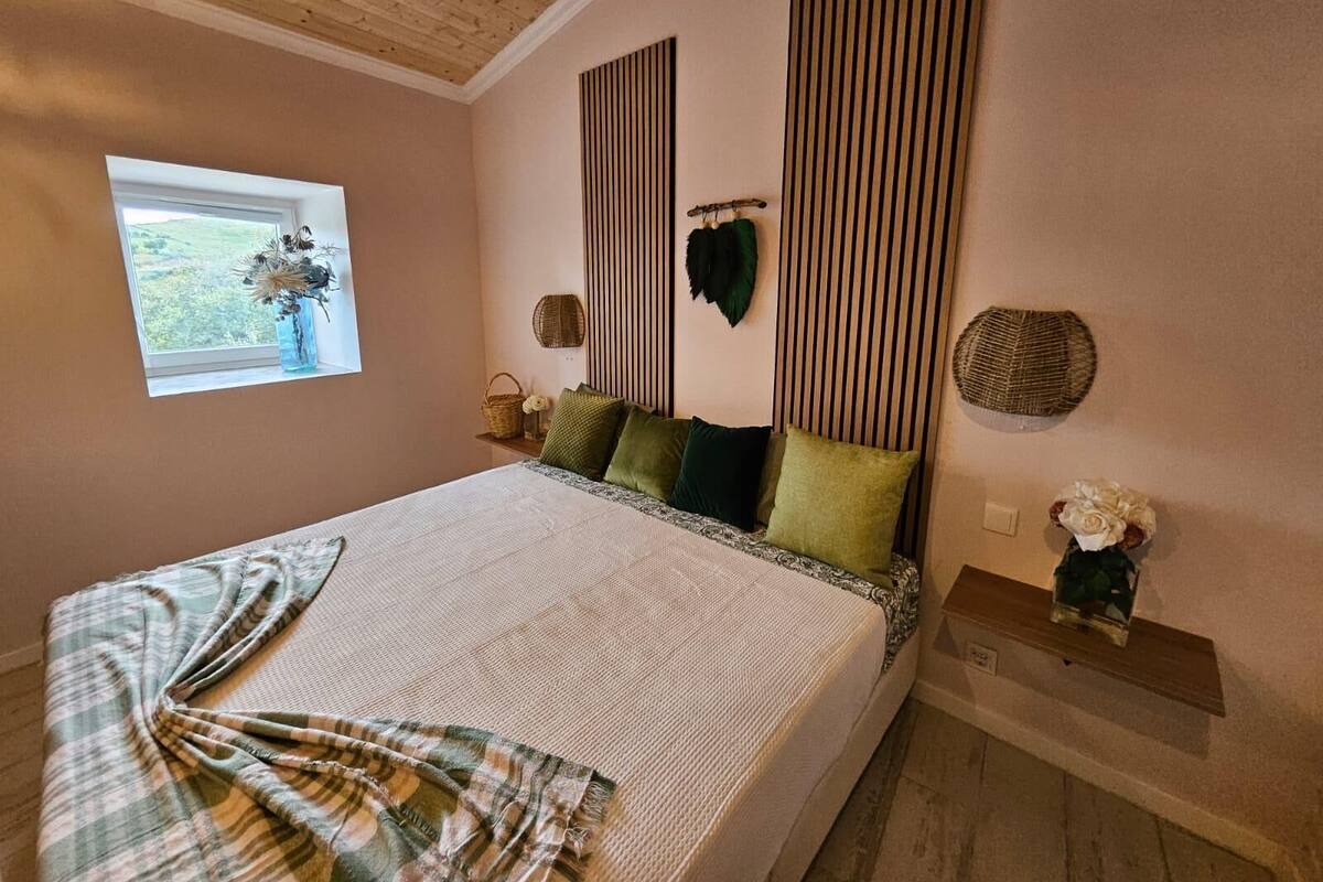 A bedroom with a bed, a window with a view, a bedside table, and a wall-mounted shelf.