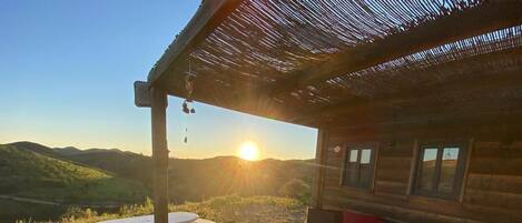 A wooden cabin with a deck, a bed with red and white pillows, and a view of the sunset over a hilly landscape.