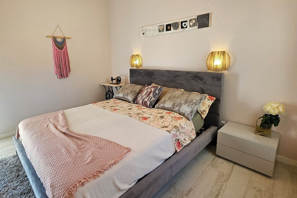 A bedroom with a wooden ceiling, a bed with floral bedding, two bedside lamps, and a small nightstand with a vase of flowers.