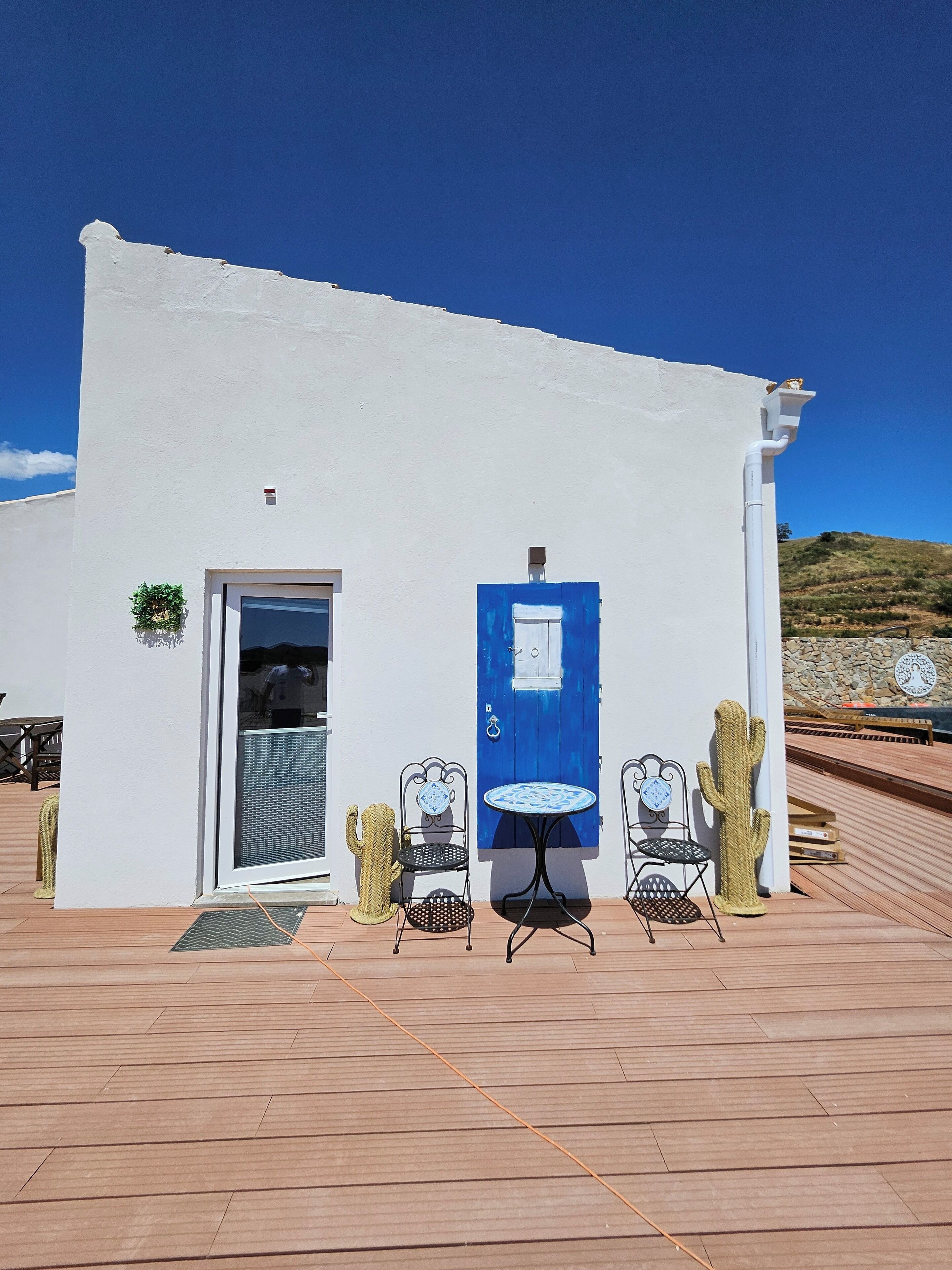 Un edificio blanco con puerta azul, dos mesas y sillas al aire libre, y una escultura de cactus.