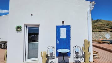 Un edificio blanco con puerta azul, dos mesas y sillas al aire libre, y una escultura de cactus.