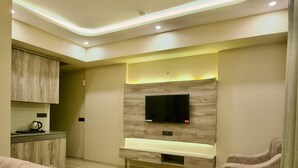 A modern living room with a sofa, a TV mounted on a wooden wall, and a kitchen area with cabinets and a coffee maker. - Villa Vistosa Gomti Nagar (Lucknow)