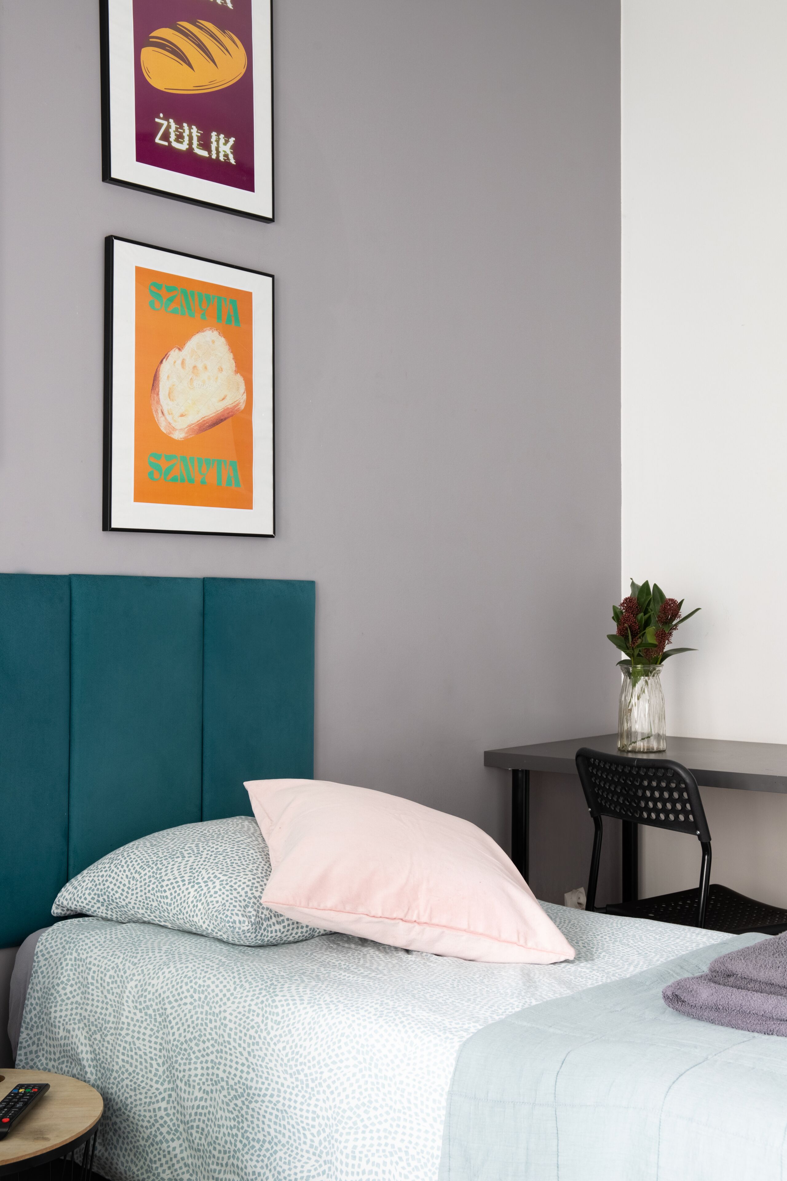 A bedroom with a teal headboard, a bed with patterned bedding, a pink pillow, a framed poster on the wall, a small table with a vase, and a chair.