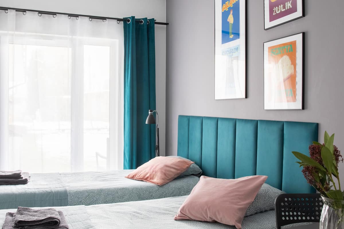 A modern bedroom with a teal headboard, a bed with a patterned bedspread, a bedside table with a vase, and two framed pictures on the wall.