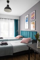 A modern bedroom with a teal headboard, a bed with a patterned bedspread, a bedside table with a vase, and two framed pictures on the wall.