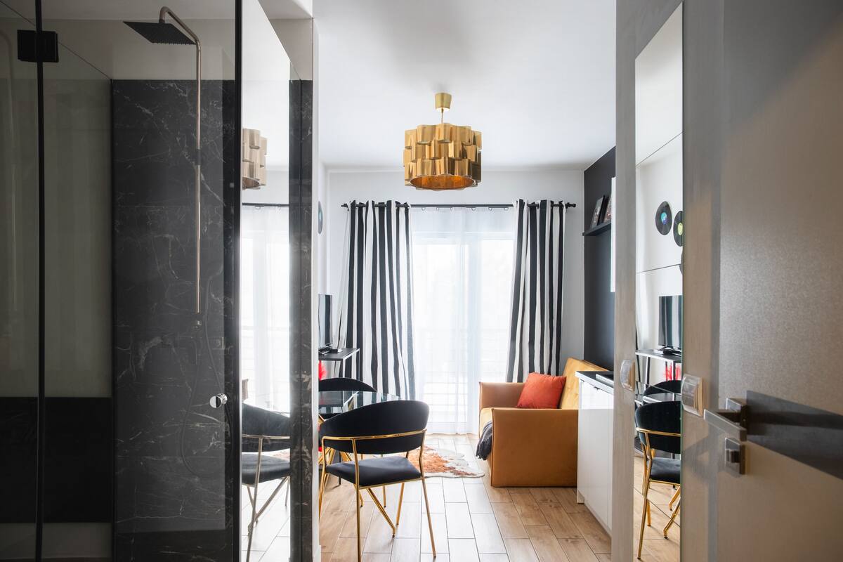 A modern interior with a glass door leading to a bathroom, a dining area with striped curtains, and a living space with a sofa and chairs.