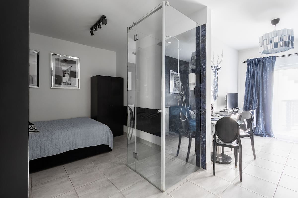 A modern room with a bed, a glass shower enclosure, a dining area with a chair, and a desk.