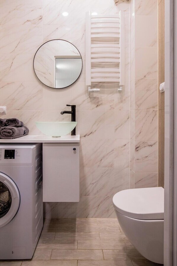 Un baño pequeño con salpicadero de mármol, un espejo redondo, un lavabo blanco, un perchero, una lavadora y un inodoro.