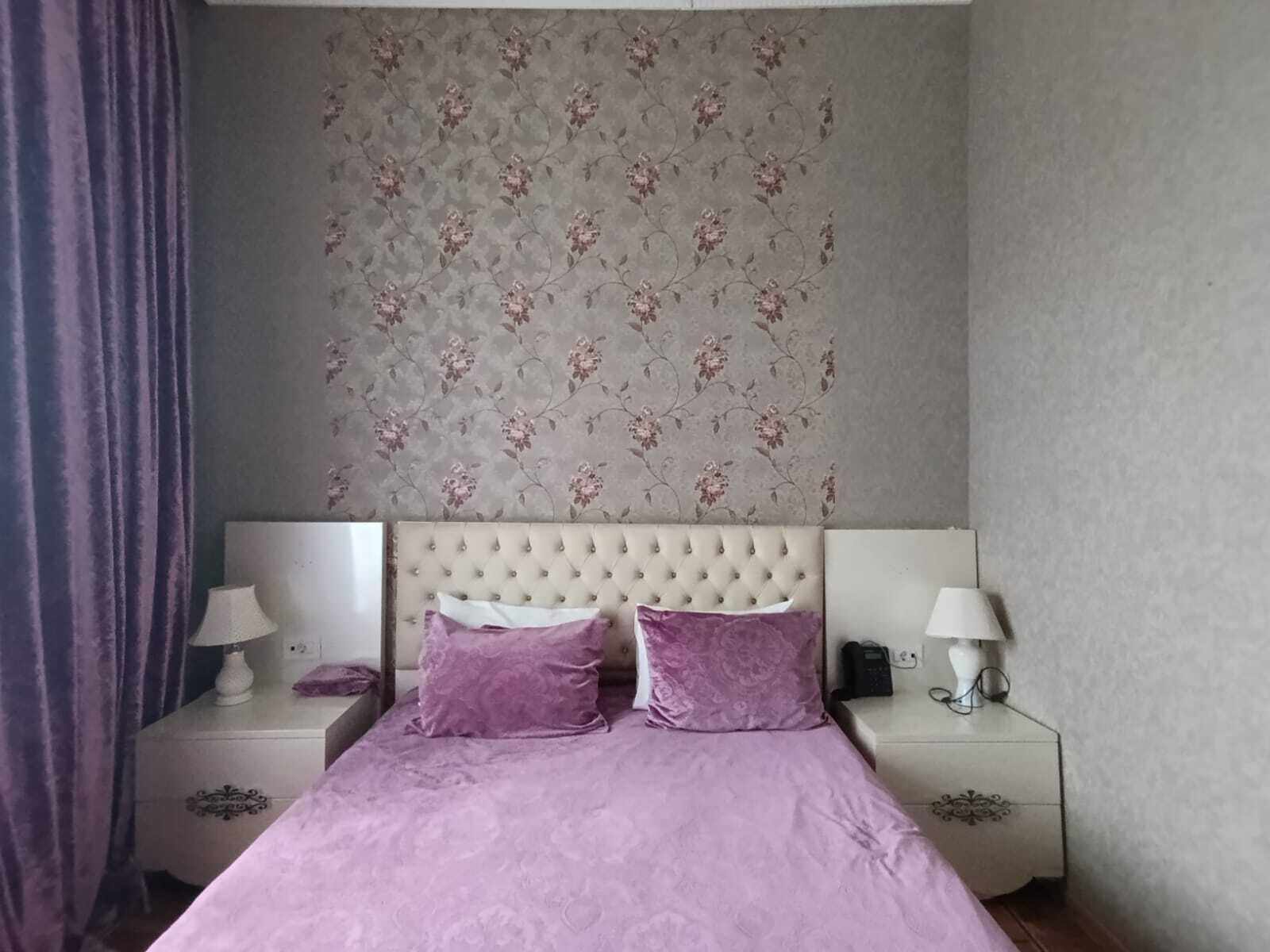 Een slaapkamer met een gestoffeerd hoofdeinde, twee nachtkastjes met lampen en bloemenbehang.
