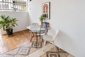 A small, enclosed patio area with a glass table and chairs, a potted plant, a framed artwork, and a tiled floor.