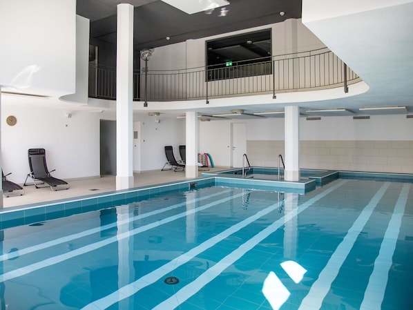 Una piscina interior con carriles claramente delimitados, zona de hamacas y un balcón con vistas a la piscina.