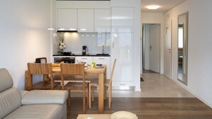 Ett modernt vardagsrum med en soffa, ett matbord i trä och stolar. I bakgrunden syns ett kök med vita skåp och en matplats.