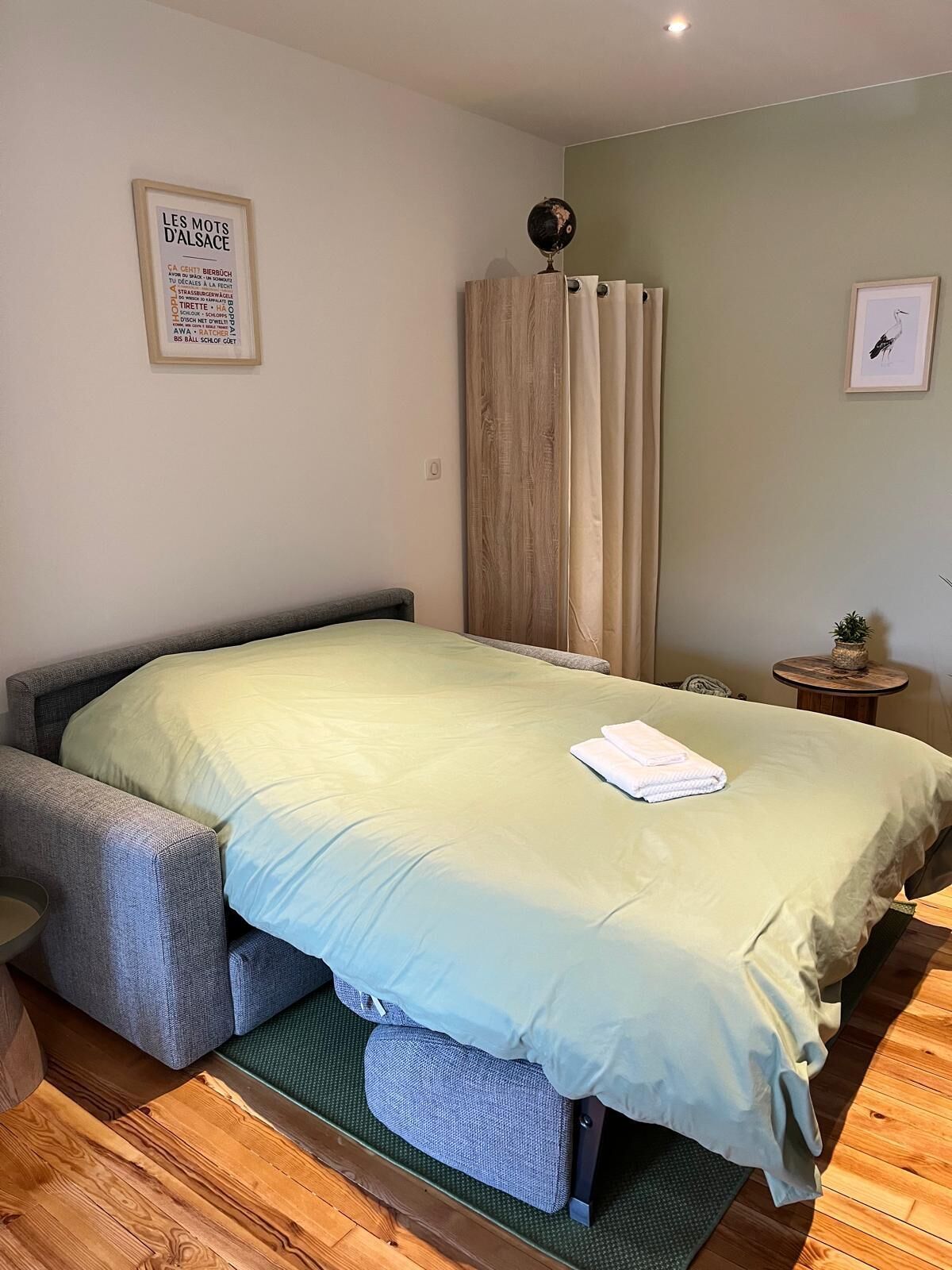A bedroom with a bed, a small bedside table, a wall-mounted lamp, and a framed poster.