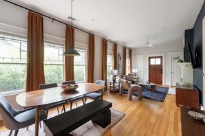 A spacious living room with a wooden dining table, comfortable seating, large windows with curtains, and a kitchen area with a refrigerator and cabinets.