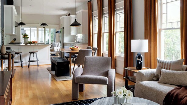 A modern living room with a dining area, large windows with curtains, and a kitchen in the background.