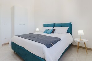 Een keurig opgemaakt bed met een blauwgroen hoofdbord, witte beddengoed en twee bedlampjes op een klein rond tafeltje.