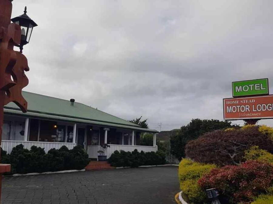 A motel with a green roof and a porch, surrounded by well-maintained bushes and a wooden signboard.