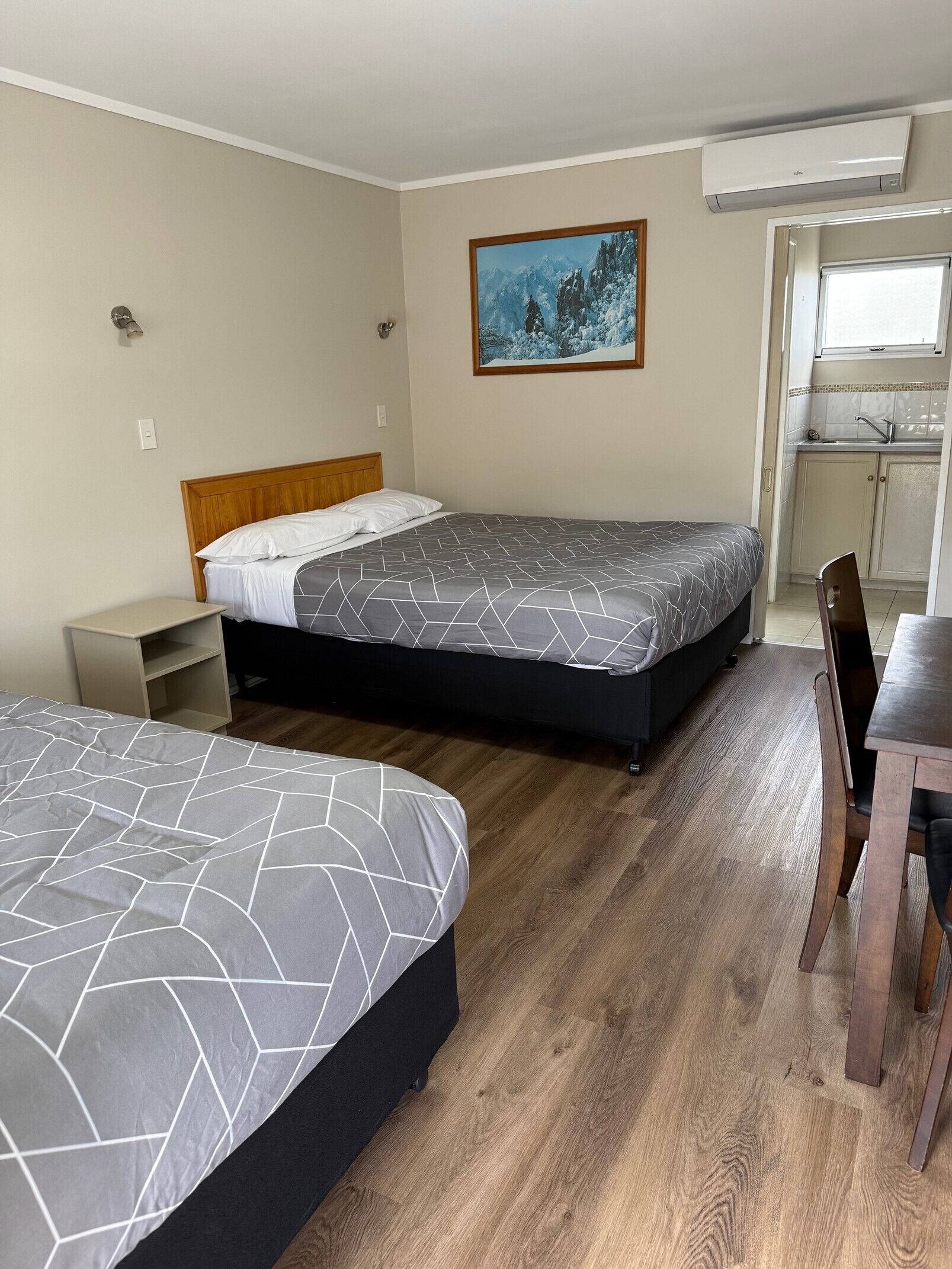 A hotel room with two beds, a small table, a chair, a painting, and a kitchenette.