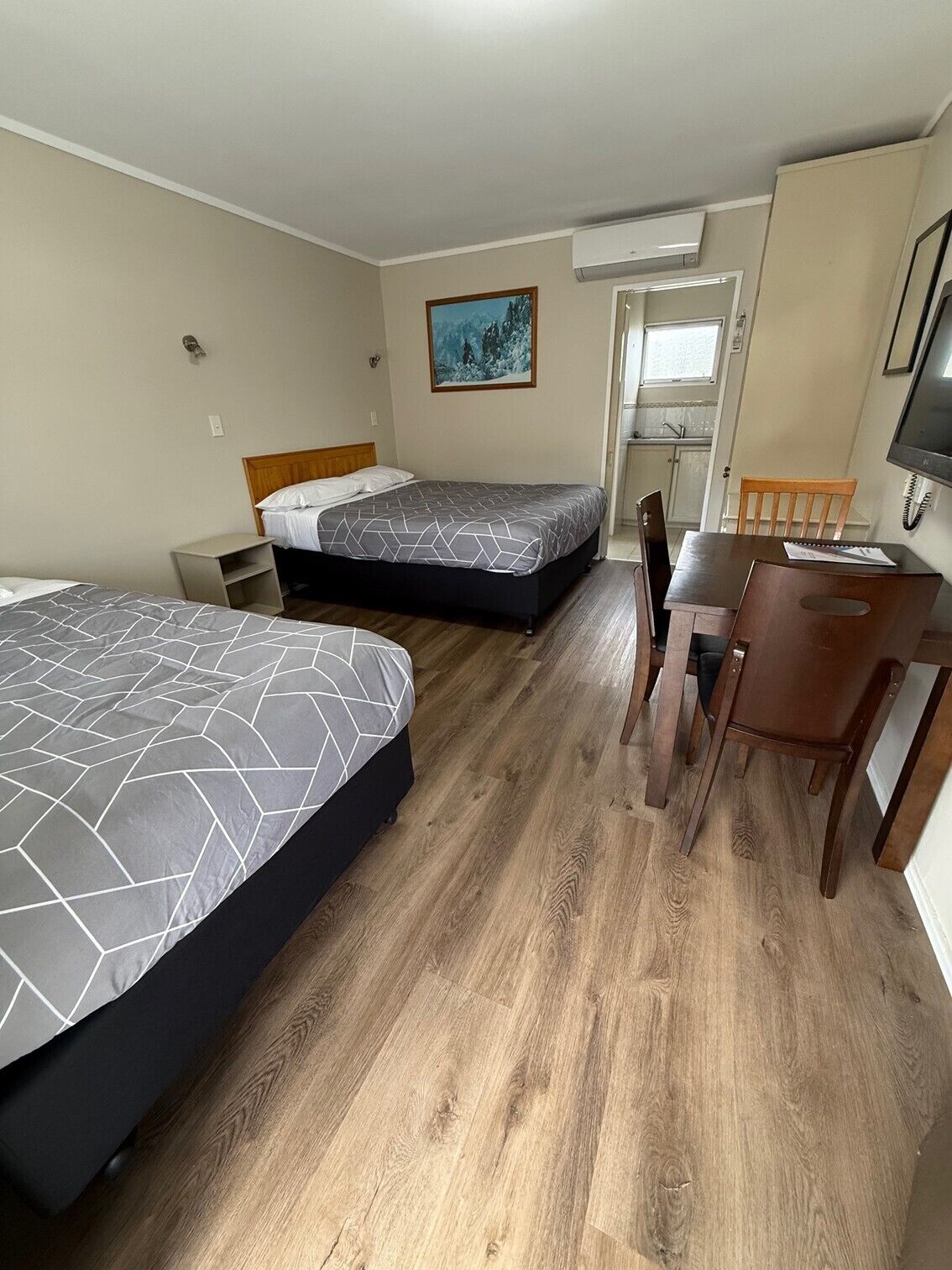 A hotel room with two beds, a wooden floor, a desk with a chair, a TV, and a painting on the wall.