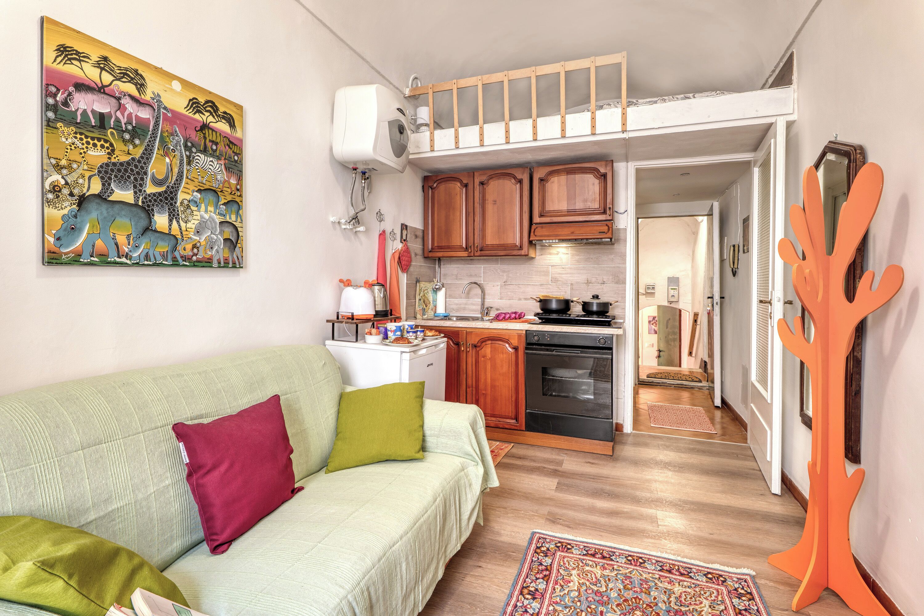A compact living space with a kitchenette, a sofa with cushions, a colorful rug, and a wall-mounted painting.