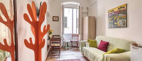 A compact living space with a sofa, a dining table, chairs, a patterned rug, a painting, and a view through a doorway.
