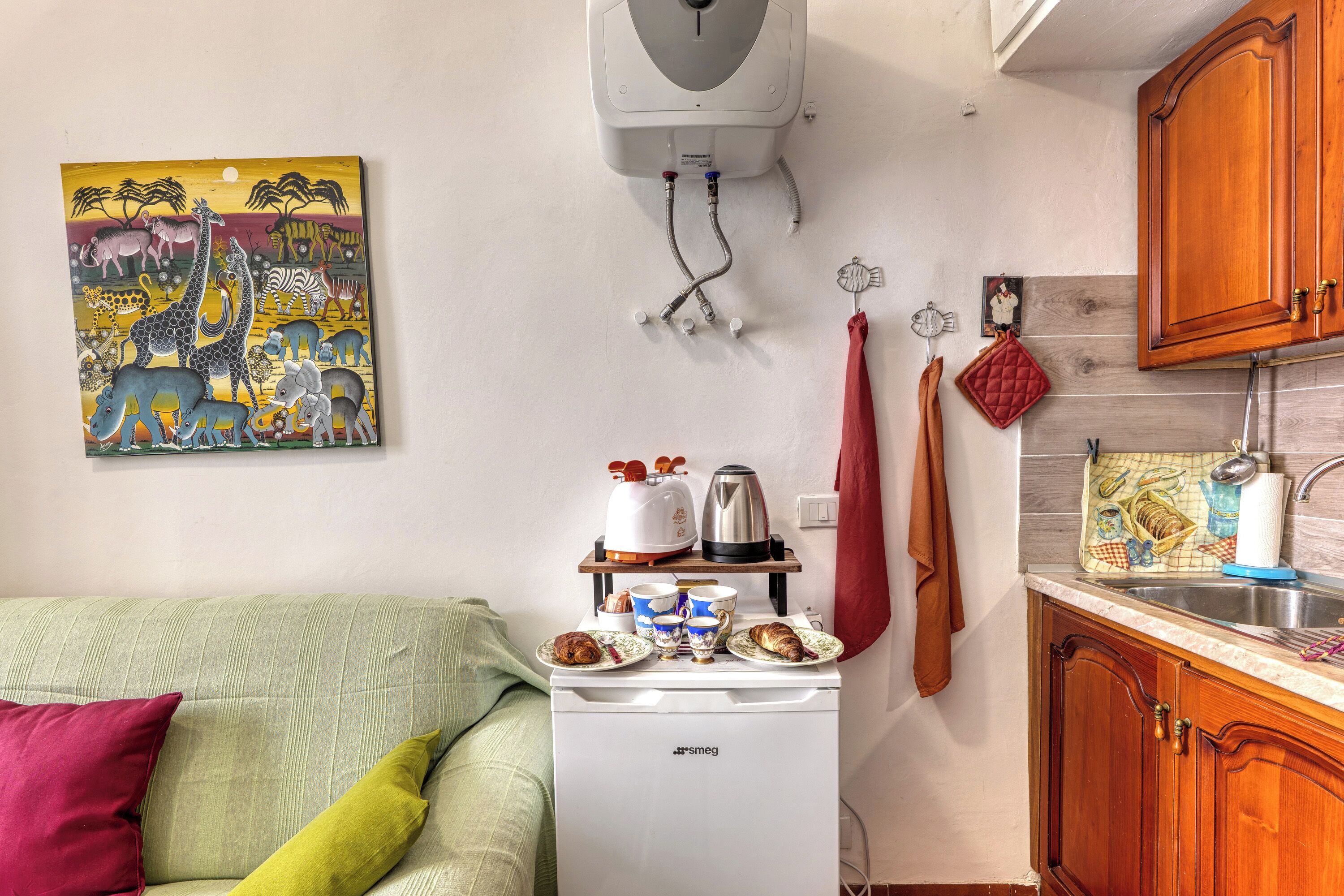 A kitchenette with a coffee maker, kettle, and cups, a small refrigerator, and a painting of animals on the wall.