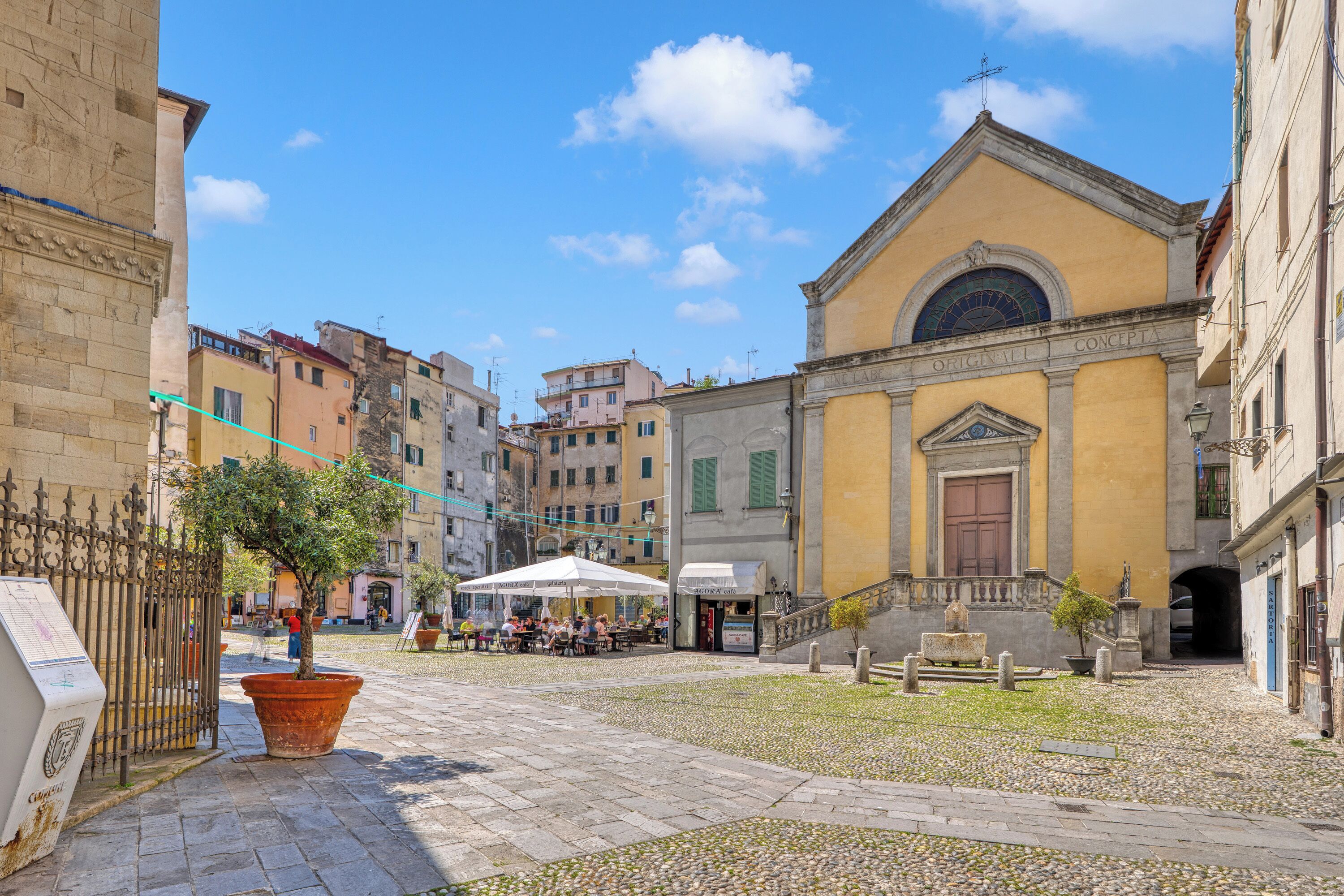 A courtyard with a building featuring a large arched window and a door, surrounded by cobblestone pavement and a few outdoor seating areas.