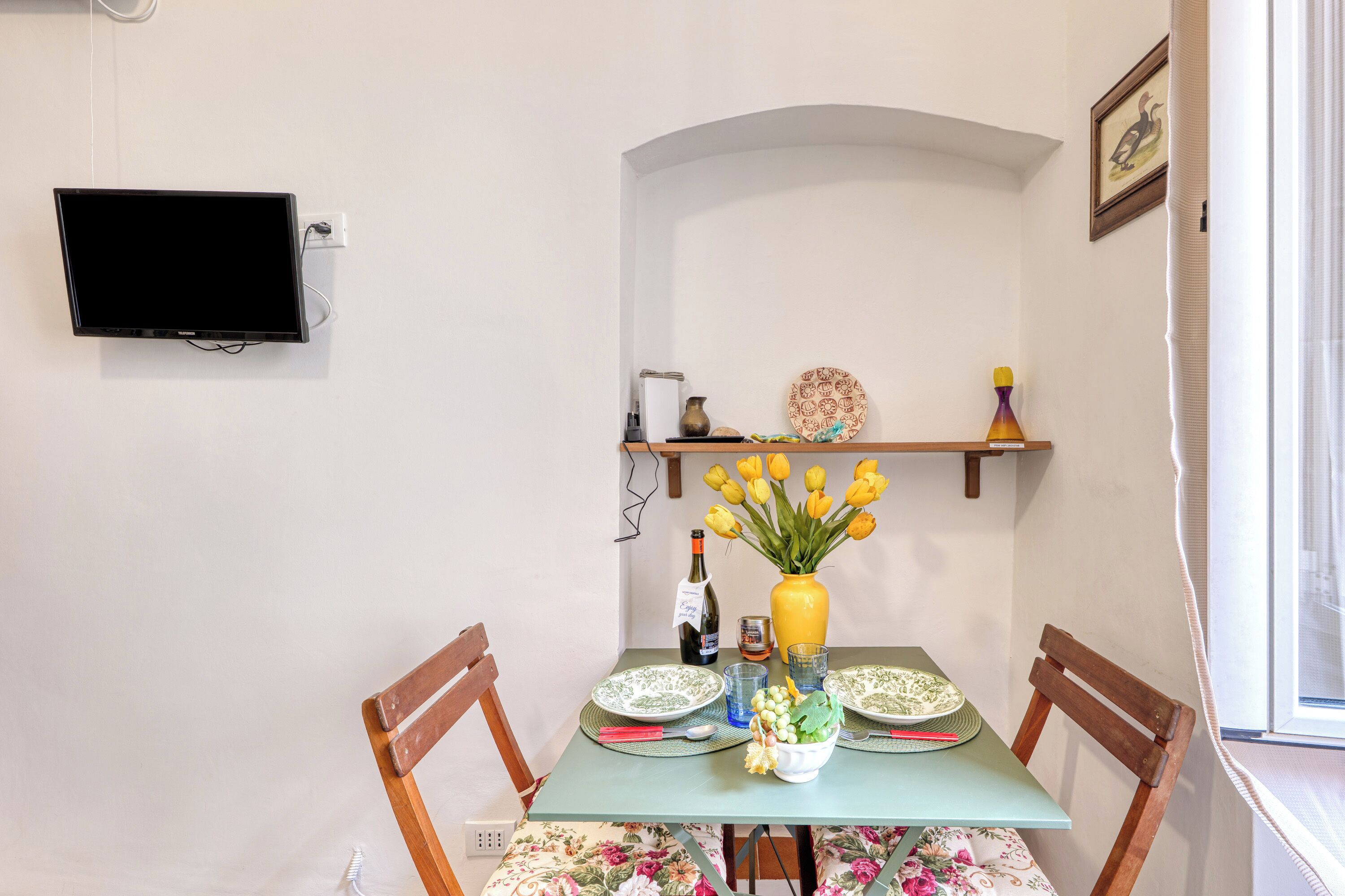 A dining area with a table set for two, a flat-screen TV mounted on the wall, and a window with curtains.