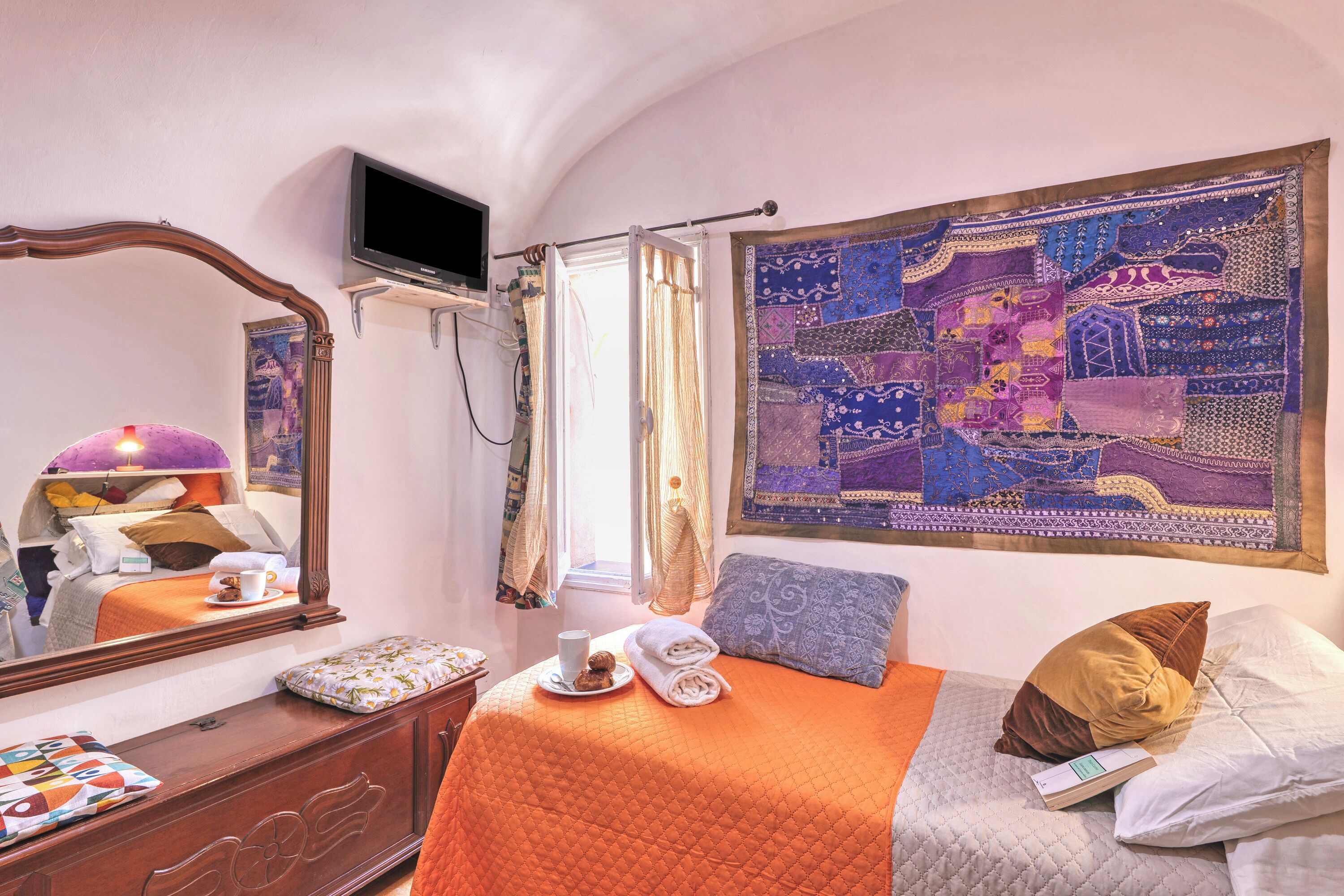 Una camera da letto con un letto, una televisione, una finestra con tende, un arazzo e un comodino con una tazza e un libro.