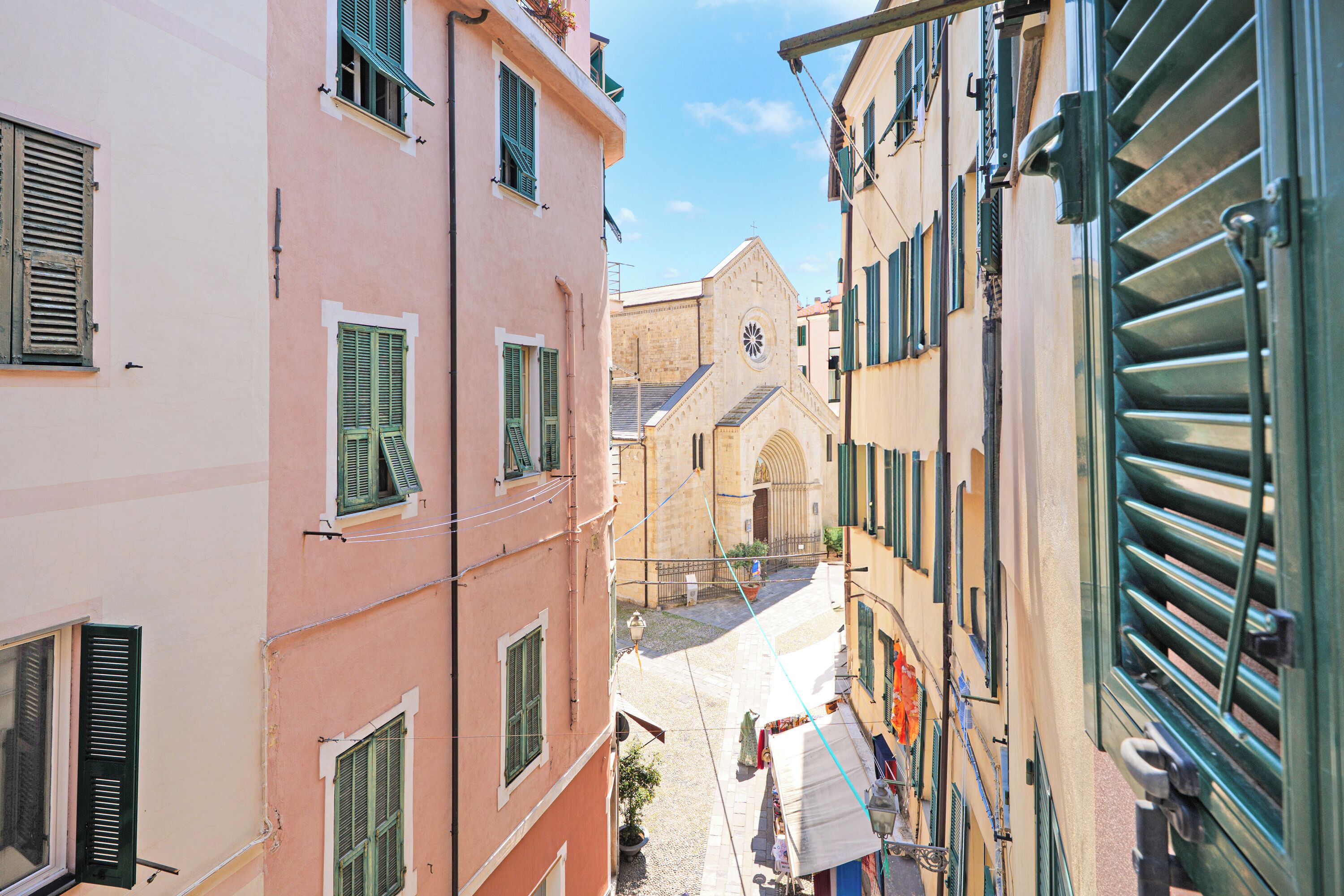 A narrow street between buildings with shutters, a church with a bell tower, and a clear sky.