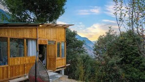 A wooden cabin with large windows, a hanging chair, and a view of the surrounding forest and mountains. - Ruhaani Cottages And Cafe (Banjar)