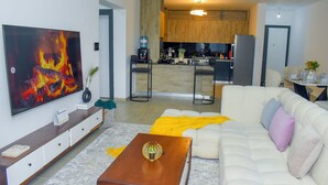 A modern living room with a large flat-screen TV, a sofa, a coffee table, and a kitchen in the background.