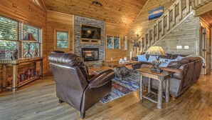 A cozy cabin interior with a fireplace, leather recliner, wooden dining table, and a staircase.