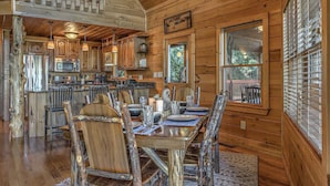 A rustic cabin interior with a wooden dining table set for four, surrounded by chairs with a natural finish. A kitchen area with wooden cabinets and a central island is visible.